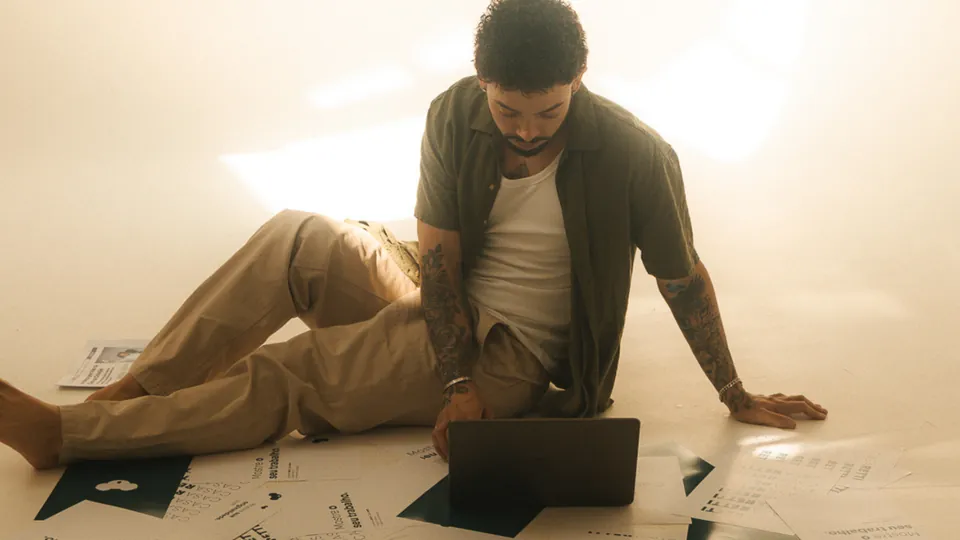 Jon sentado no chão do estúdio, trabalhando no notebook entre papéis de planejamento